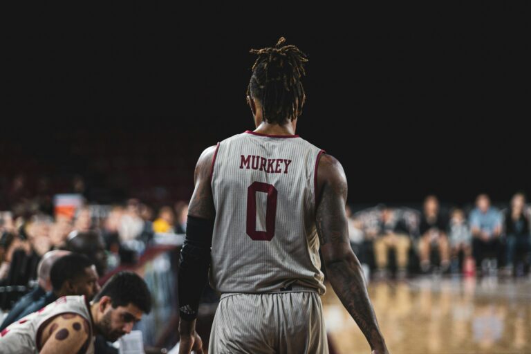 a college basketball player in a shite and burgundy jersey standing on the sidelines with his back to the camera
